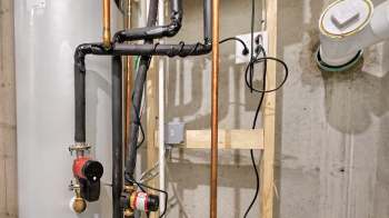 Pipes and valves connected to a water heater in a basement, with electrical components mounted on a wooden frame.