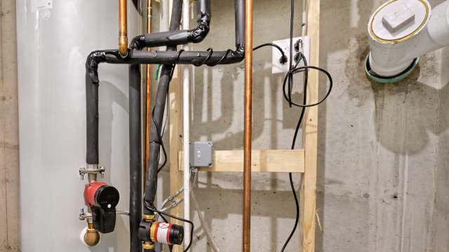 Pipes and valves connected to a water heater in a basement, with electrical components mounted on a wooden frame.