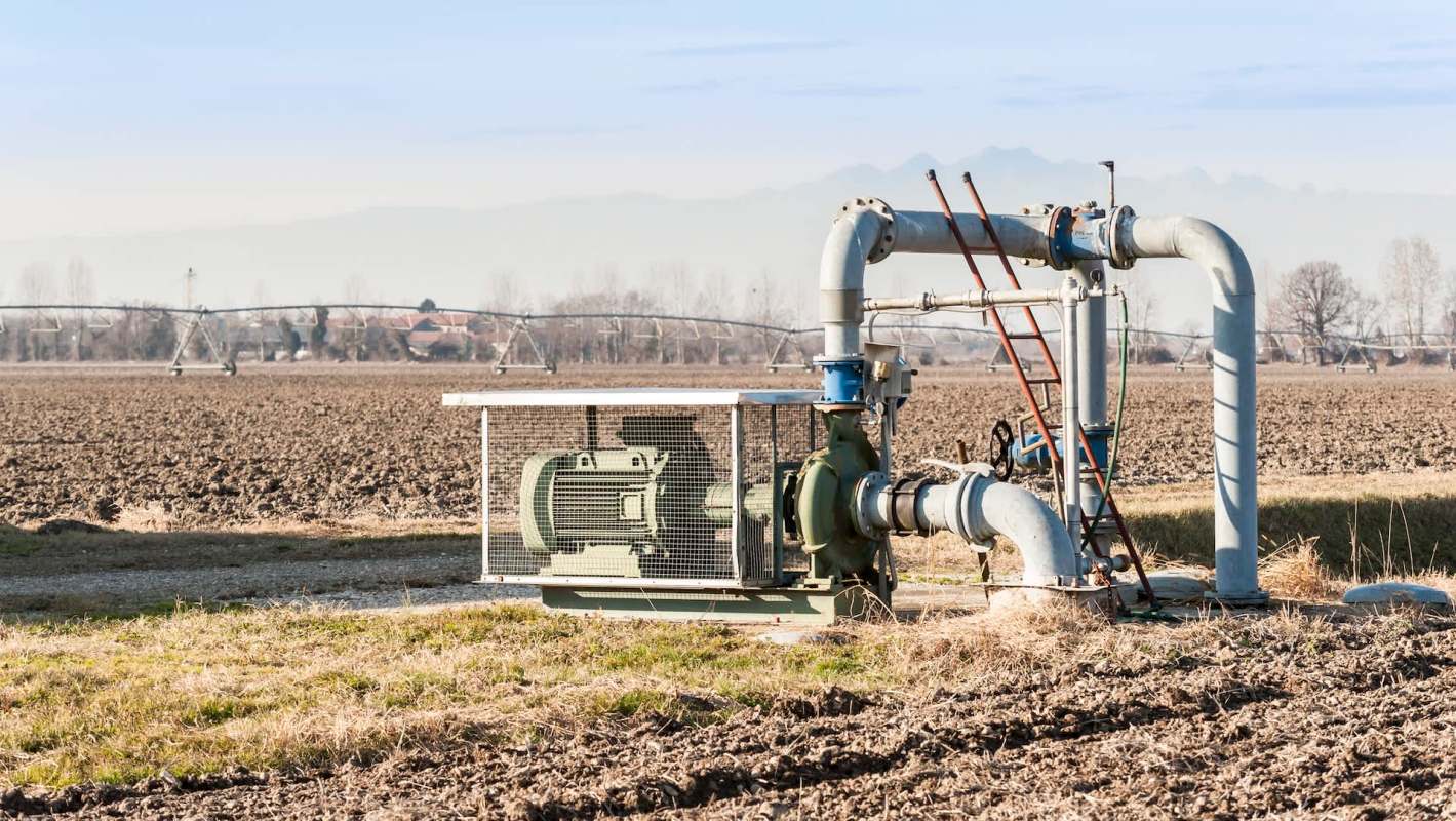A water pump system with pipes is installed on a dry agricultural field under a clear sky.
