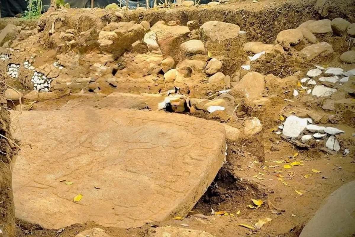 An archaeological site featuring exposed stone structures and carvings on a large rock surrounded by dirt and smaller stones.