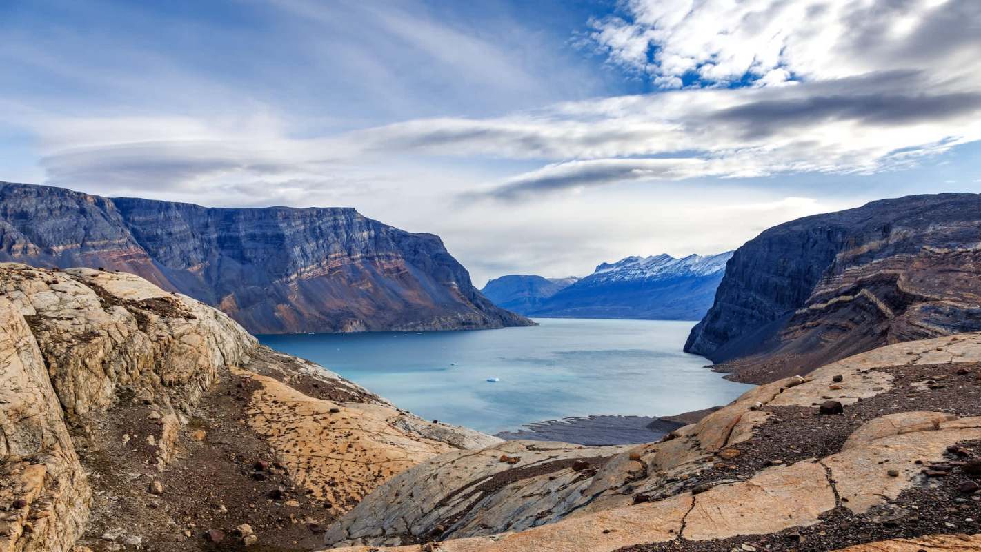 A panoramic view of a fjord surrounded by rocky cliffs and snow-capped mountains under a partly cloudy sky.