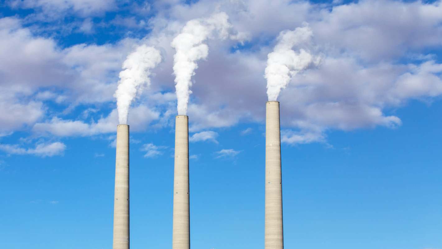 Three industrial chimneys emitting white smoke against a blue sky with scattered clouds.