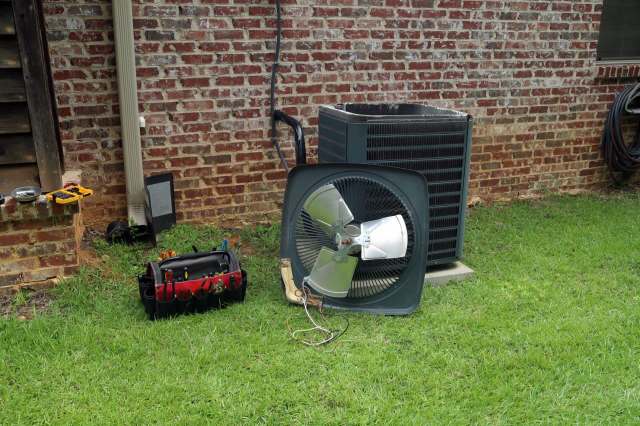 An air conditioning unit with a detached fan and a toolbox on grass near a brick wall.
