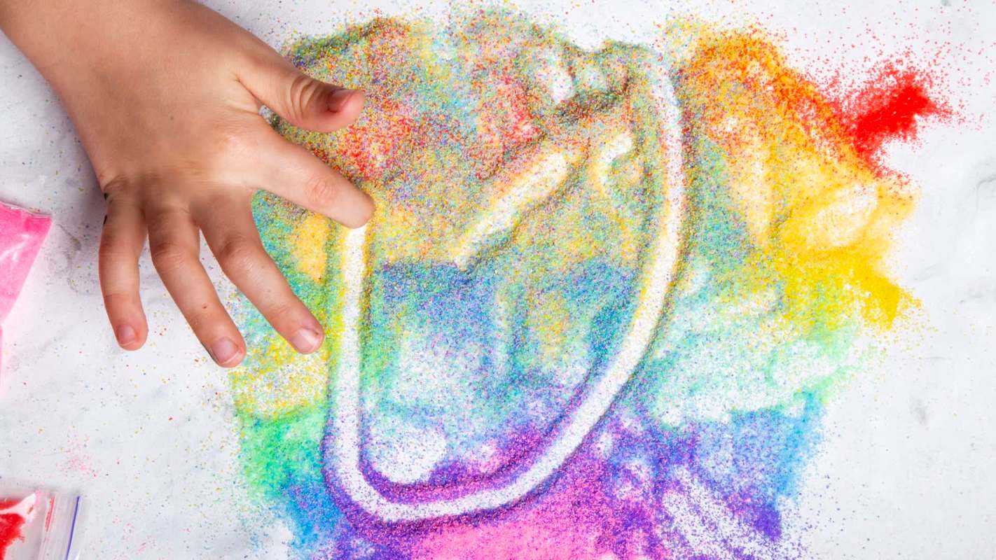 A child's hand creates colorful patterns with bright, glittery sand on a white surface.