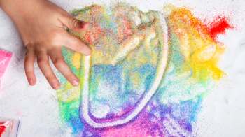 A child's hand creates colorful patterns with bright, glittery sand on a white surface.
