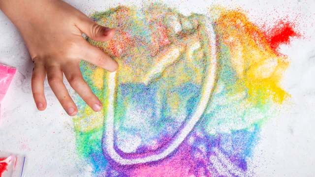 A child's hand creates colorful patterns with bright, glittery sand on a white surface.