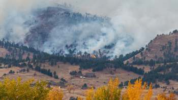 A wildfire burns in the mountains, with smoke rising and flames visible among the trees and hills.