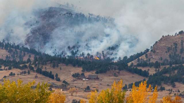 A wildfire burns in the mountains, with smoke rising and flames visible among the trees and hills.