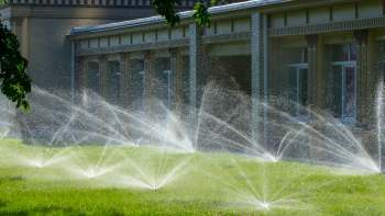 Sprinklers are watering a lush green lawn near a brick building on a sunny day.