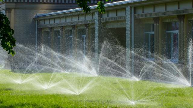 Sprinklers are watering a lush green lawn near a brick building on a sunny day.