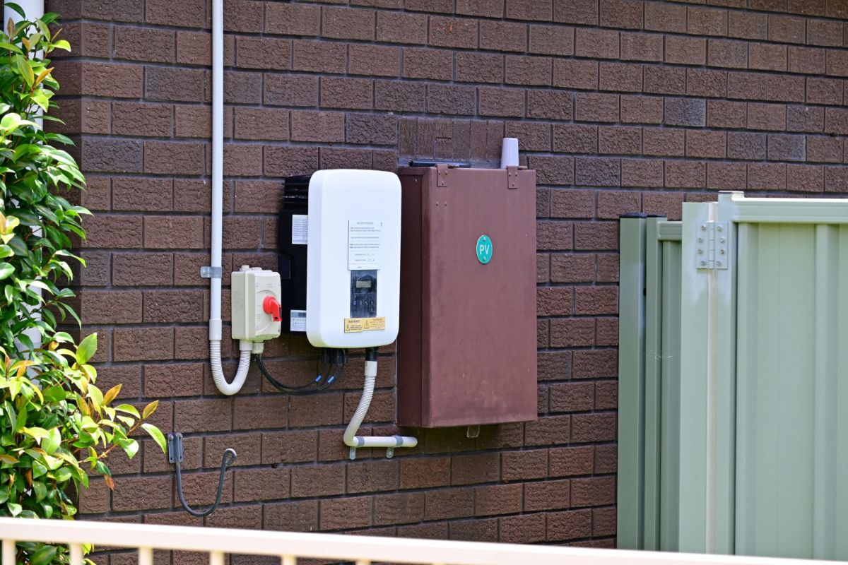 An exterior wall features a white electrical box and a brown panel, surrounded by green foliage and a metal fence.