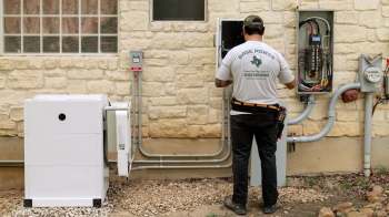 A technician works on electrical equipment outside a building, including an inverter and a service panel.