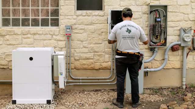 A technician works on electrical equipment outside a building, including an inverter and a service panel.