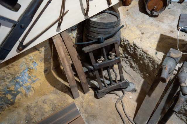 An old wooden press with metal components, surrounded by various tools and wooden planks in a rustic setting.