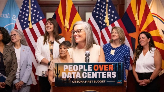 Arizona Gov. Katie Hobbs stands behind a podium displaying "People Over Data Centers" with flags in the background.