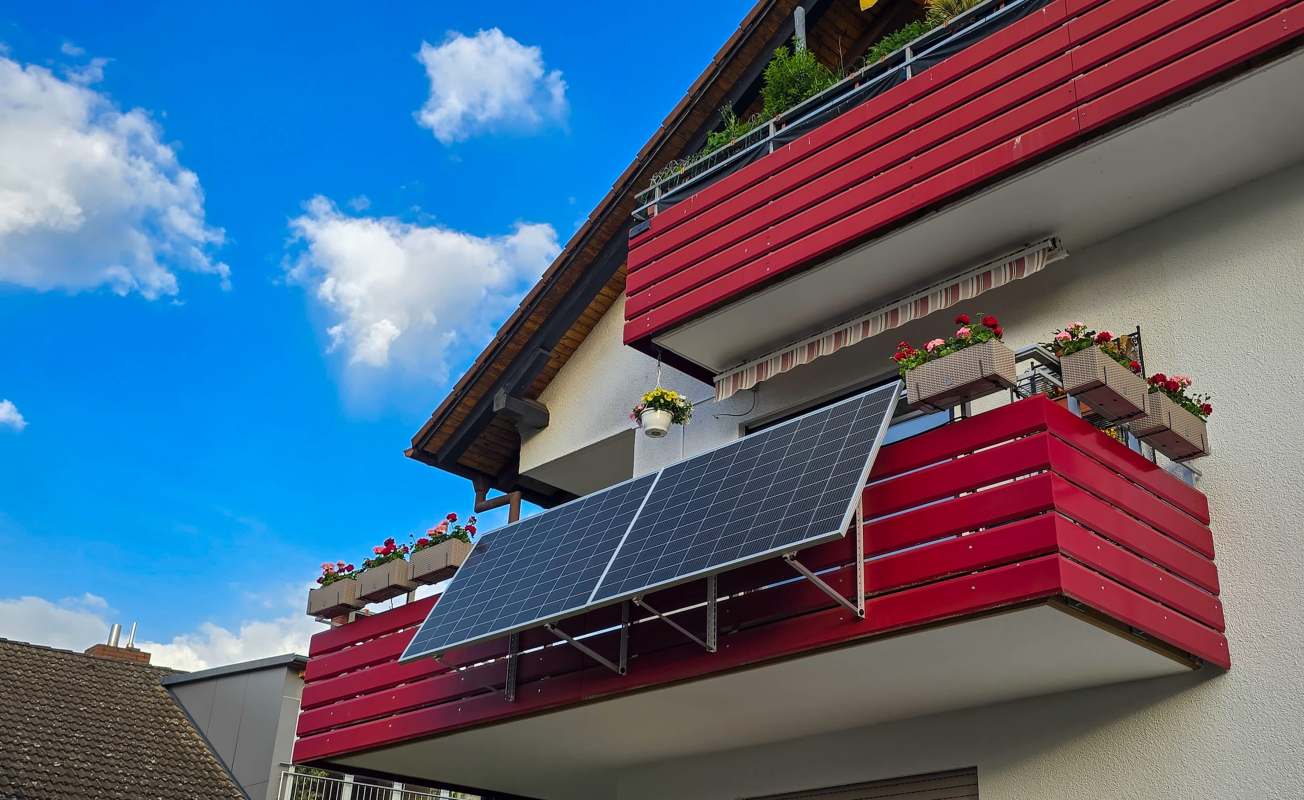 A modern balcony with red wooden panels, solar panels, and flower pots under a blue sky filled with clouds.