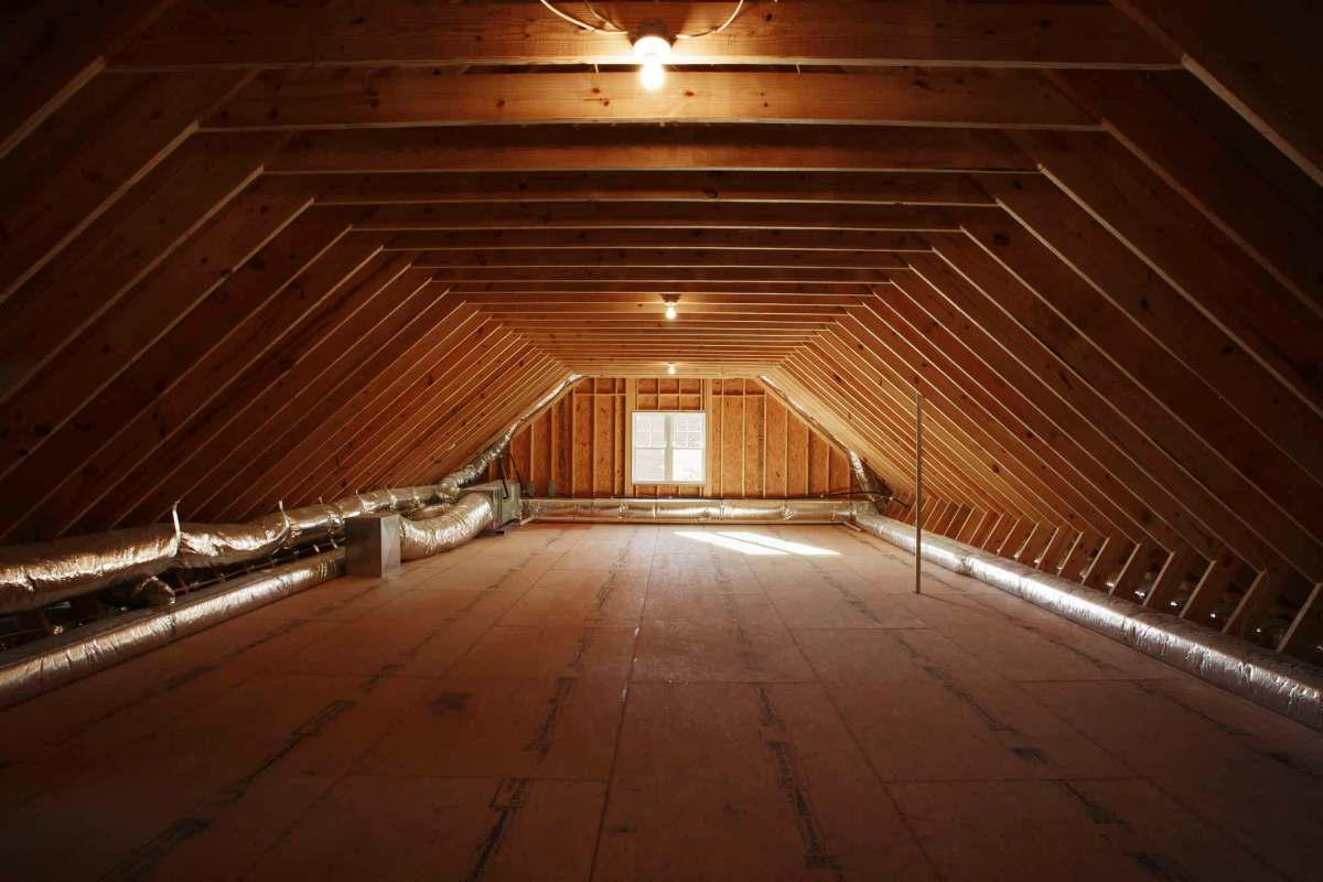 An empty attic with slanted wooden beams, exposed insulation, and a window allowing natural light.
