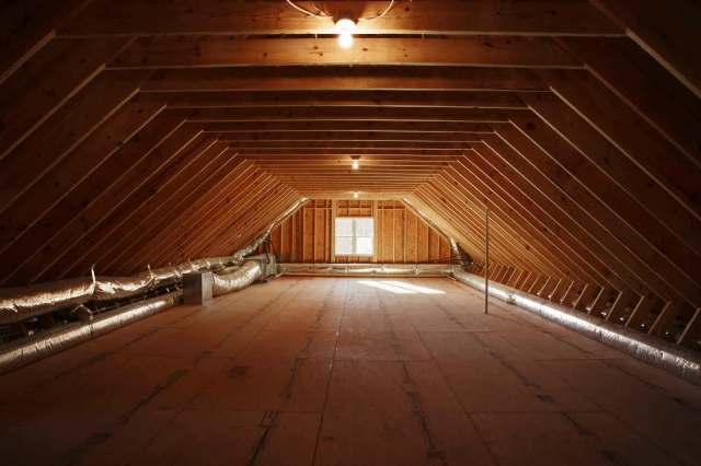 An empty attic with slanted wooden beams, exposed insulation, and a window allowing natural light.