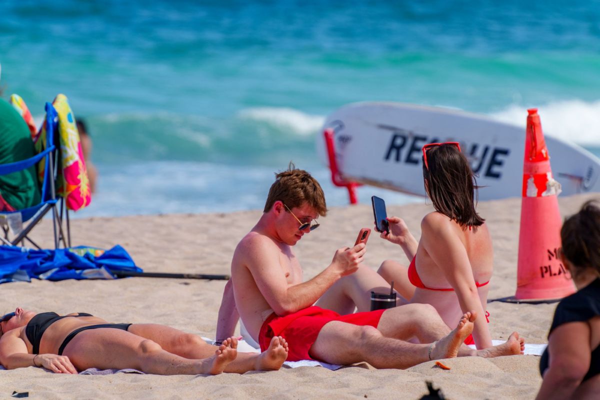 A group of young adults on the beach, looking at their phones.