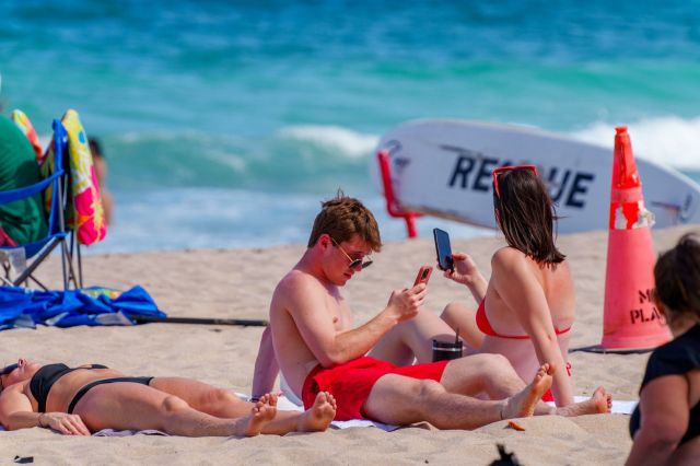 A group of young adults on the beach, looking at their phones.