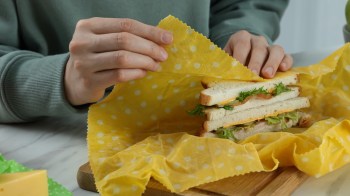 A hand gently unwraps a sandwich wrapped in yellow polka dot beeswax paper on a wooden cutting board.