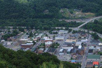An aerial view of a small town with green hills, showing various buildings, streets, and parked cars.