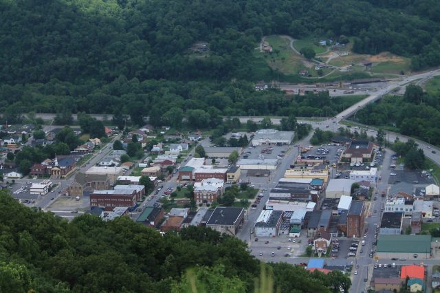 An aerial view of a small town with green hills, showing various buildings, streets, and parked cars.
