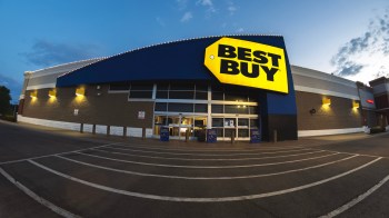 A Best Buy store exterior illuminated at dusk, showcasing a prominent sign and parking lot.