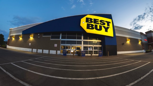 A Best Buy store exterior illuminated at dusk, showcasing a prominent sign and parking lot.