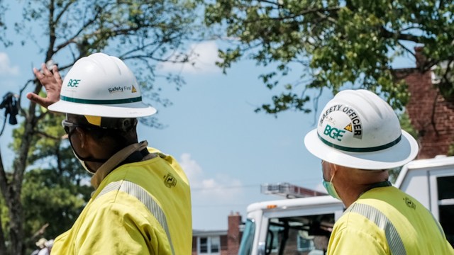 Two utility workers in safety gear discuss while standing outdoors under a clear blue sky.