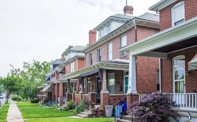 A row of red-brick houses with front porches and greenery lining the sidewalk on a cloudy day.
