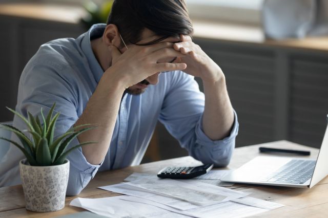 A stressed man with his hands on his head sits at a table cluttered with papers and a calculator.