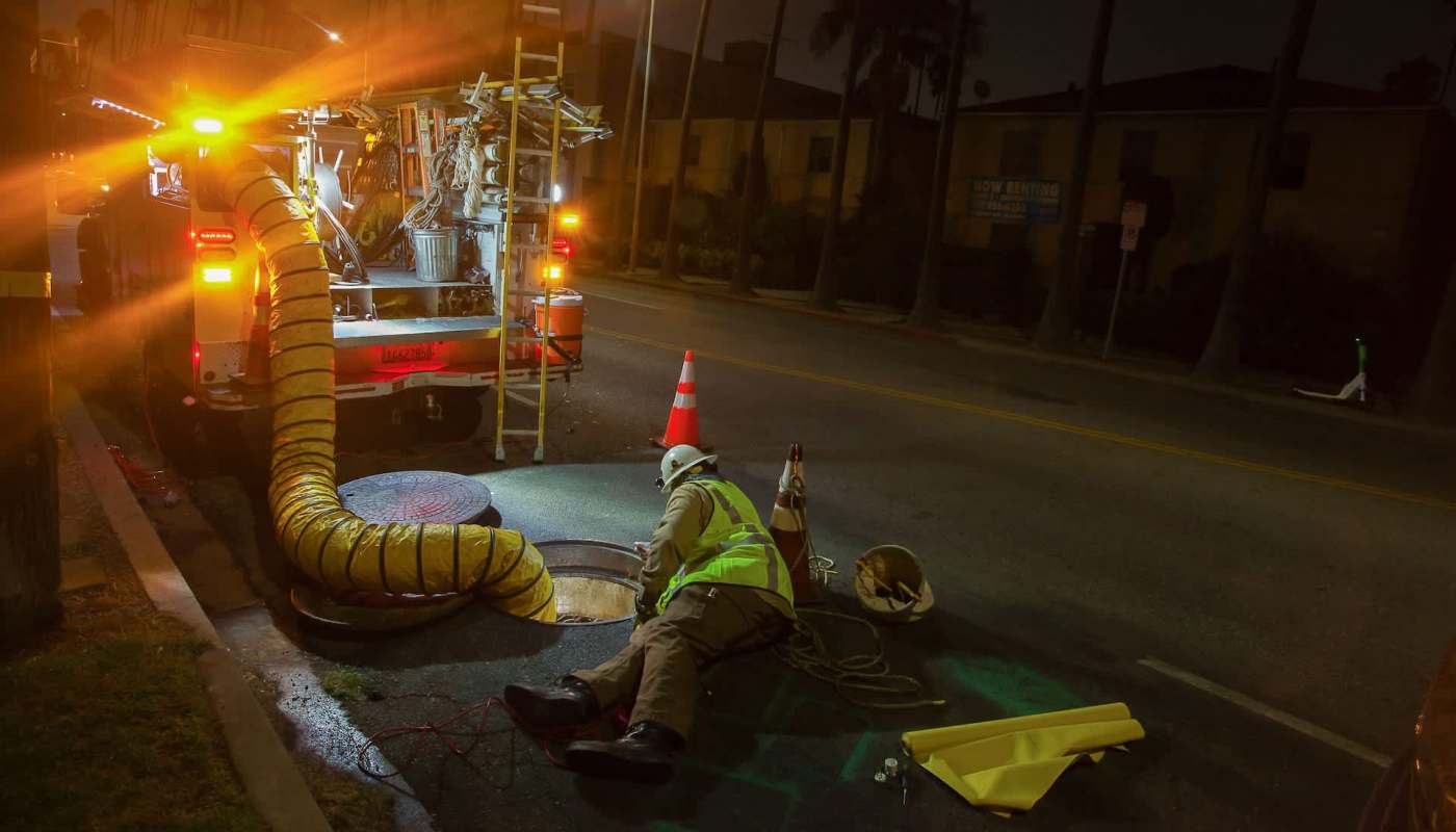 A worker in a reflective jacket repairs a manhole with a large hose connected to a service truck at night.