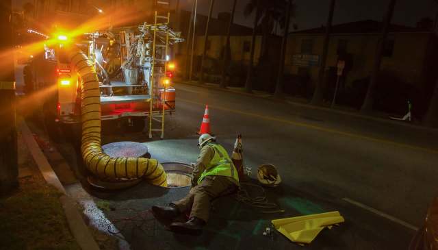 A worker in a reflective jacket repairs a manhole with a large hose connected to a service truck at night.