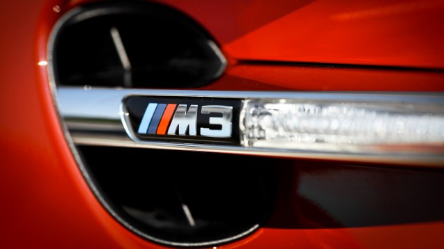 A close-up of a BMW M3 badge on an orange car with a sleek design and reflected light.