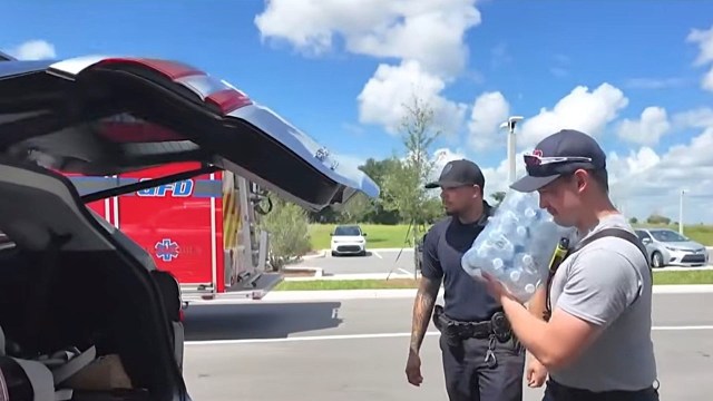 Two emergency responders are outdoors, one holding a pack of bottled water while another stands nearby.