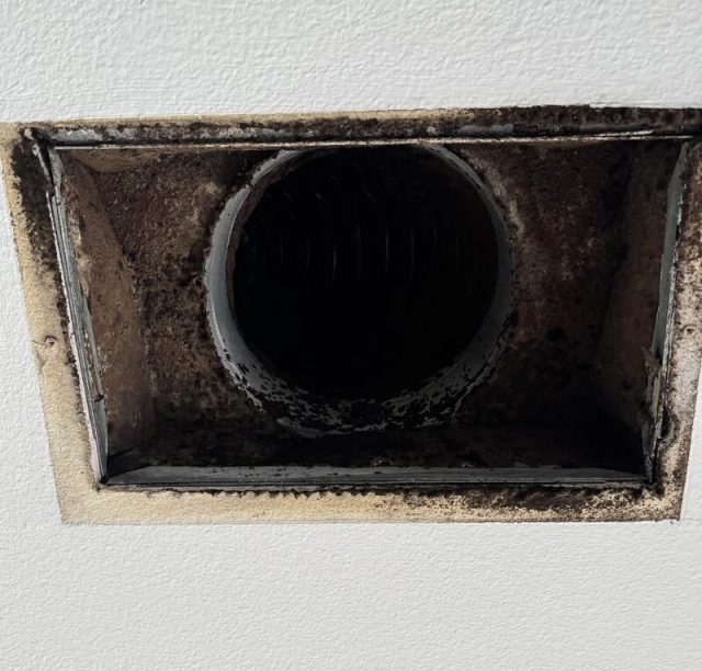 A person wearing blue gloves removes a dirty air vent cover from a wall in a light blue room.