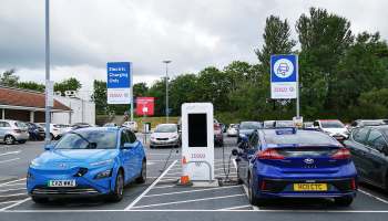 Two blue electric vehicles parked at a charging station in a Tesco supermarket lot, with signs indicating charging availability.