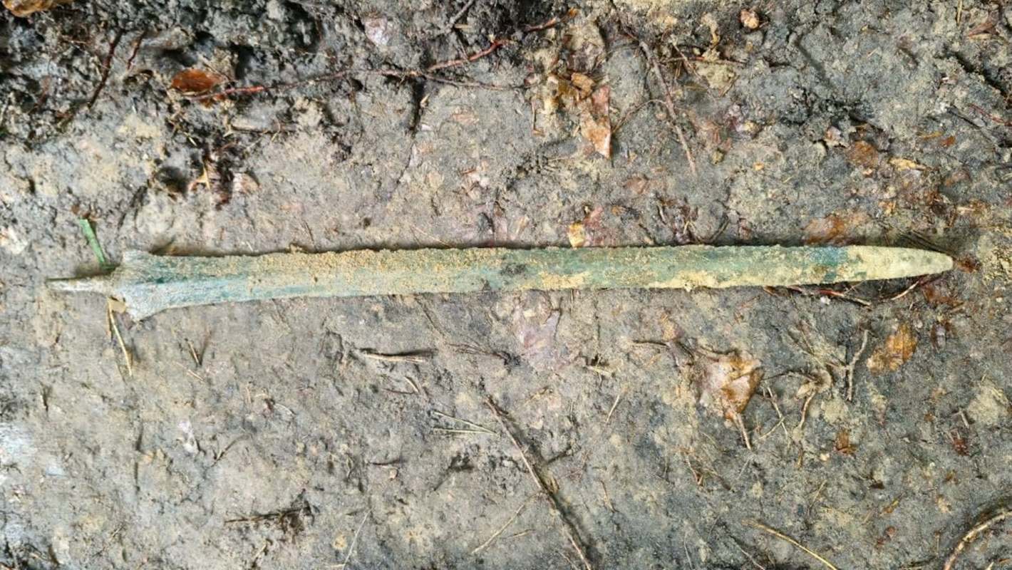 An ancient, weathered bronze sword lies partially buried in muddy ground surrounded by leaves and debris.