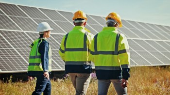 Three workers in safety gear walk past solar panels in a grassy field under a clear blue sky.