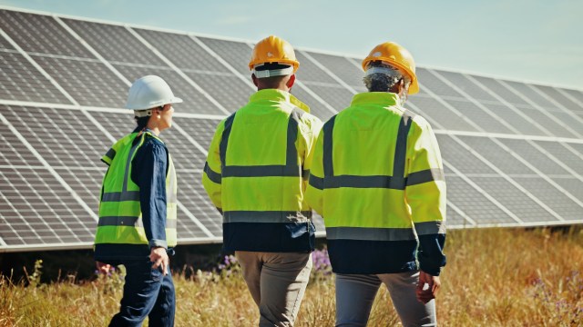 Three workers in safety gear walk past solar panels in a grassy field under a clear blue sky.
