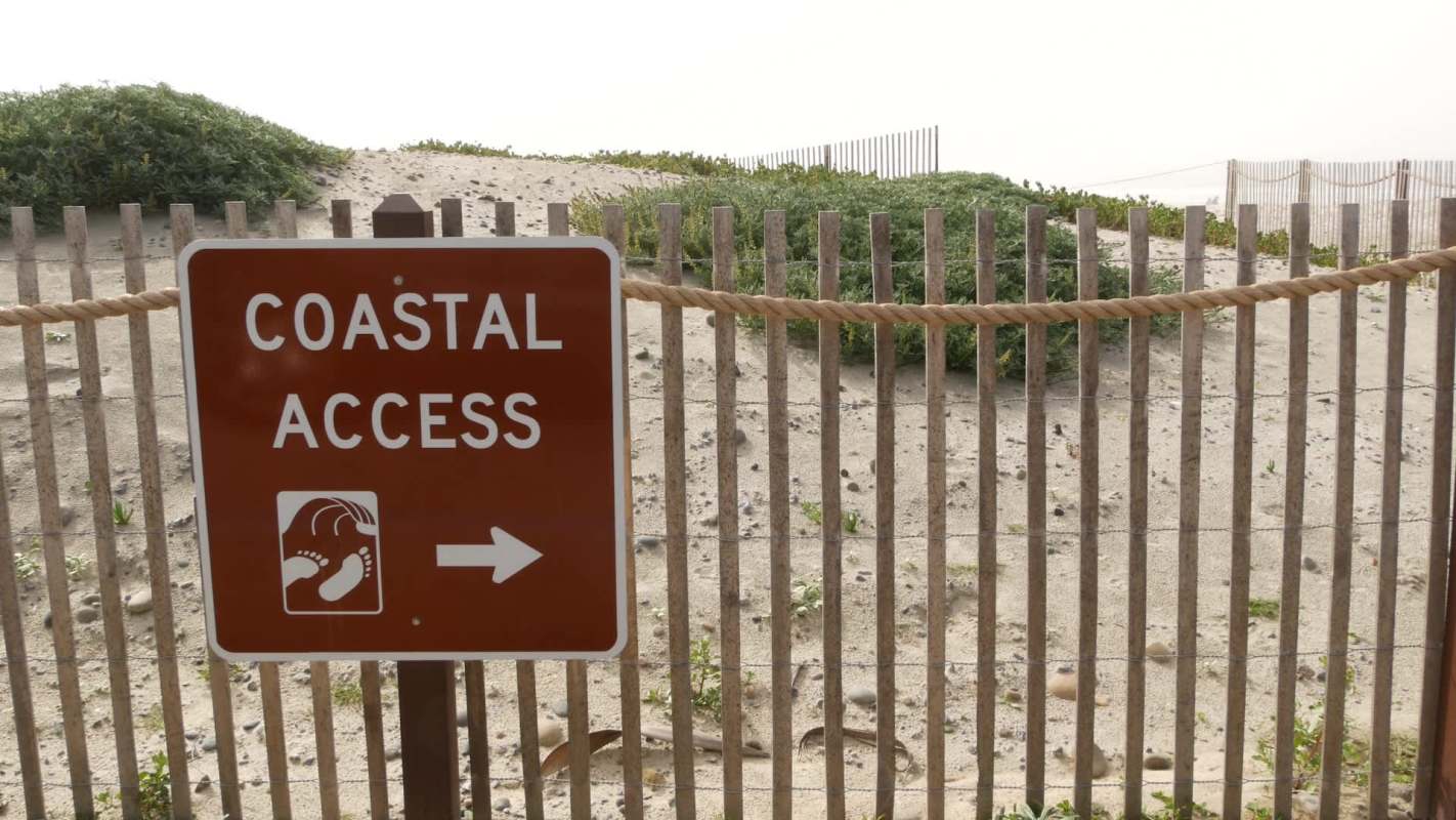 A sign indicating coastal access with footprints and an arrow pointing right, near sandy terrain and plants.