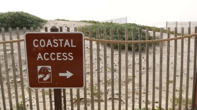 A sign indicating coastal access with footprints and an arrow pointing right, near sandy terrain and plants.