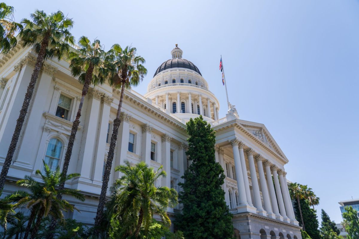 A close-up view of a grand building (Caifornia state capitol) with a dome, palm trees, and a clear blue sky.