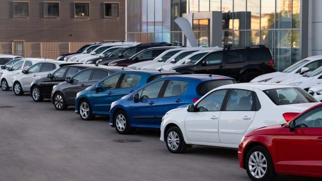 A row of various cars in a dealership parking lot, featuring blue, white, black, and red vehicles.