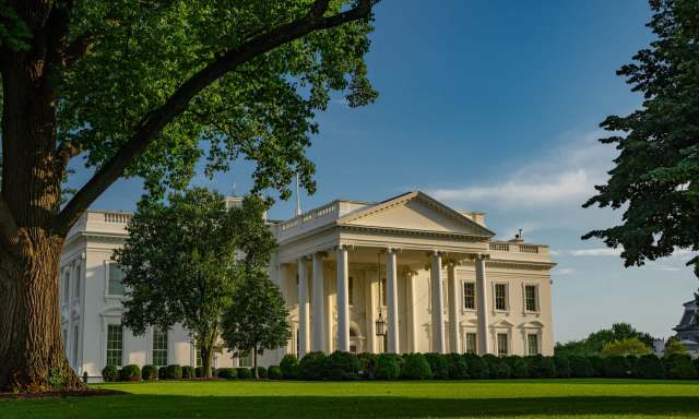 The White House is shown surrounded by lush green trees and manicured lawns under a clear blue sky.