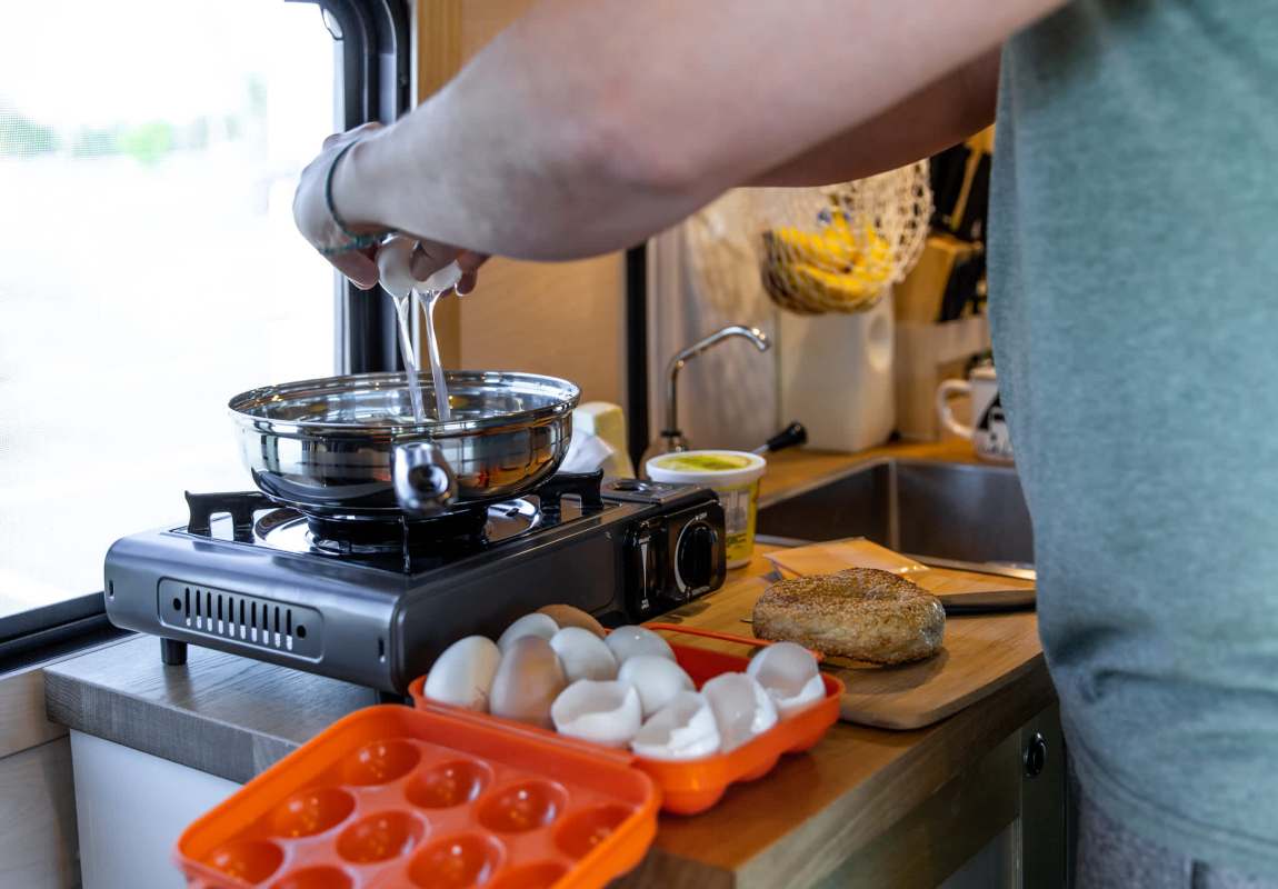 A person cracks eggs into a frying pan on a portable stove in a cozy kitchen setting.
