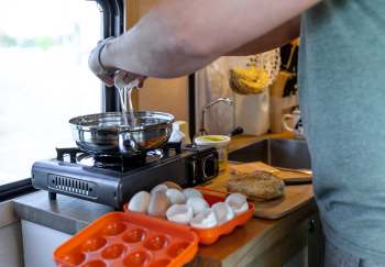 A person cracks eggs into a frying pan on a portable stove in a cozy kitchen setting.