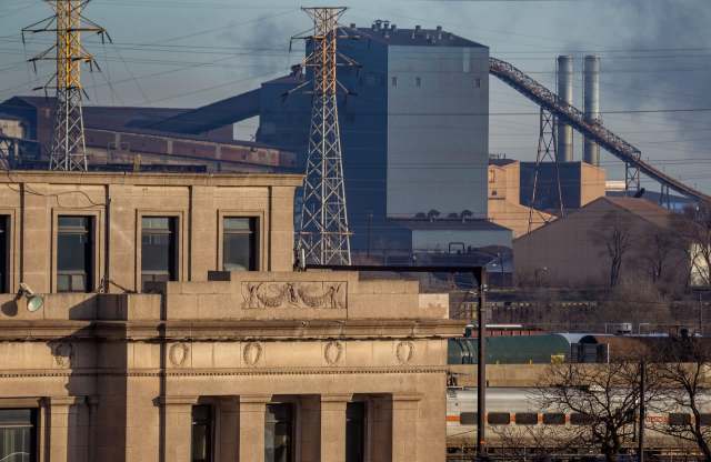 A view of a steel mill with power lines and smoke in the background, against a clear sky.