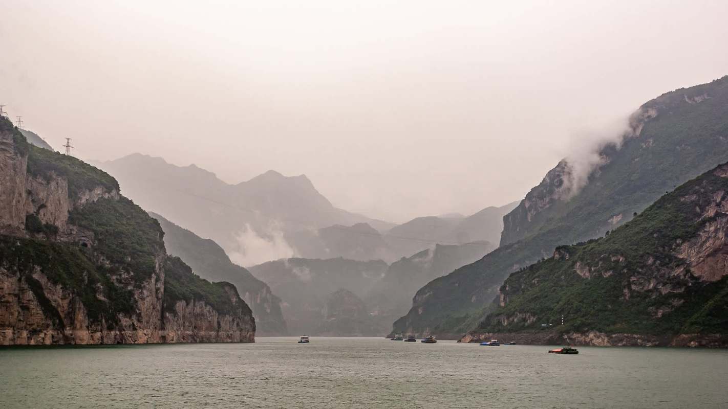 A misty landscape of mountains with a river, featuring boats amidst lush green cliffs.